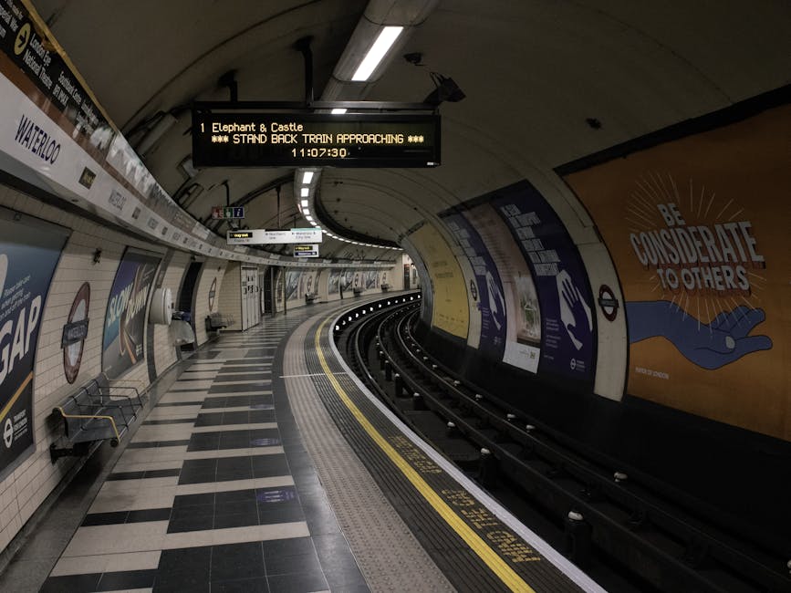 An empty underground train station platform with curved tiled walls and ceiling, illuminated by overhead fluorescent lights. The platform features a black and white checkered tile pattern on the floor, with a yellow tactile strip along the edge for safety. On the left side, there are benches and advertising posters attached to the tiled wall, including a prominent one with the message 'BE CONSIDERATE TO OTHERS' featuring an illustration of a blue hand on a yellow background. Above the platform, a digital display board indicates the approaching train to 'Elephant & Castle' via Waterloo, scheduled to arrive at 11:07:30. The curved tracks run along the right side of the platform, with a dark tunnel exit visible in the background. The scene has a clean, quiet appearance typical of an urban metro station, providing a neutral, detailed view relevant to infrastructure and transportation environments where waste and rubbish management services may operate within the station premises.