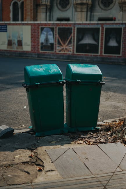 The image shows two green outdoor rubbish bins with hinged lids placed side by side on a concrete sidewalk near the curb. The bins are made of durable plastic with a smooth, slightly glossy finish, and have black handles on the lids for lifting and securing. They are positioned in front of a brick and plaster building with decorative window frames and ornate architectural details in the background, suggesting an urban environment. The surrounding area includes a paved street with asphalt and a section of worn pavement with a mix of concrete and stone slabs at the edge. Small leaves and gravel are scattered at the base of the bins, and the lighting indicates daytime with natural light casting soft shadows. The presence of the bins suggests they are part of a private waste collection or on-site clearance service, aligning with alternatives to regular municipal rubbish disposal, as represented by [COMPANY_NAME], which could offer rubbish removal services in this setting.