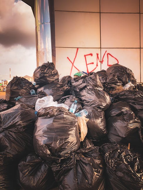 A large pile of black garbage bags filled with waste materials is stacked against a modern building with a tiled, reflective exterior. Some bags appear bulging and tied at the top, containing unknown refuse, with a few transparent plastic bottles and miscellaneous rubbish protruding from the bags. Behind the pile, there is a wall with beige square tiles, and red graffiti spelling 'XEND' can be seen spray-painted on the tiles. The scene is outdoors, on a paved surface, possibly near an urban commercial or residential area. The lighting suggests late afternoon or early evening, with a cloudy sky overhead. The image illustrates an instance of unmanaged waste accumulation that a private rubbish removal service such as [COMPANY_NAME] might manage as part of an alternative, on-site waste clearance operation.