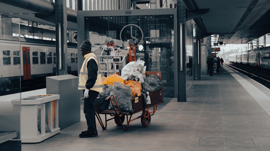 A person wearing a dark jacket, black trousers, white gloves, and a grey beanie stands next to a large red-handled trolley filled with various bags and loose rubbish, including black plastic bin bags, orange bags, and white plastic waste sacks. The trolley is positioned on a paved platform within a modern train station, with train tracks visible to the right and multiple train carriages, predominantly silver with red and black accents, parked alongside. Behind the individual, there is a glass-enclosed elevator shaft and contemporary station architecture featuring steel supports and glass panels. The scene is illuminated by natural daylight, creating a clear, neutral setting typical of a busy urban transport hub, where private waste collection or rubbish removal activities are discreetly occurring in the context of alternative waste handling within the station environment.