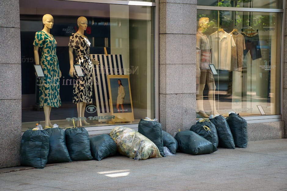 A large pile of black garbage bags filled with waste materials is stacked against a modern building with a tiled, reflective exterior. Some bags appear bulging and tied at the top, containing unknown refuse, with a few transparent plastic bottles and miscellaneous rubbish protruding from the bags. Behind the pile, there is a wall with beige square tiles, and red graffiti spelling 'XEND' can be seen spray-painted on the tiles. The scene is outdoors, on a paved surface, possibly near an urban commercial or residential area. The lighting suggests late afternoon or early evening, with a cloudy sky overhead. The image illustrates an instance of unmanaged waste accumulation that a private rubbish removal service such as [COMPANY_NAME] might manage as part of an alternative, on-site waste clearance operation.