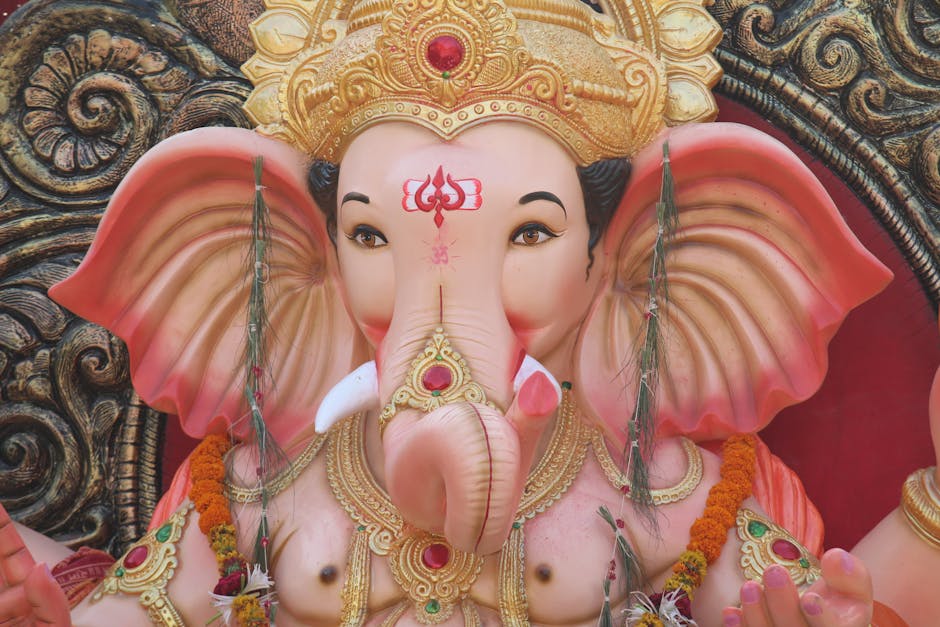 A detailed close-up of a large statue of the Hindu deity Ganesha, featuring a pinkish-orange elephant head with large, textured ears on either side. The statue's face has a smooth, polished finish with a light pink hue, and expressive dark eyes with defined eyebrows. It wears an ornate golden crown adorned with red gemstones and intricate scrolling patterns, positioned atop headgear decorated with floral motifs. The trunk is centered, extended downward, and decorated with gold and red accents, with a gold nose ring attached to it. The deity's broad shoulders are draped with traditional jewelry, including layered gold necklaces with intricate filigree work and red gemstone embellishments, as well as strings of marigold flowers and small white flowers hanging from the neck. The background features a decorative carved wooden panel with swirling patterns painted in dark colors, providing a contrast to the vibrant and detailed figure of Ganesha. This image captures the divine and ornamental elements typical of Hindu religious statues, highlighting craftsmanship and vibrant religious symbolism in a setting that could be found in a temple, festival, or shrine area.