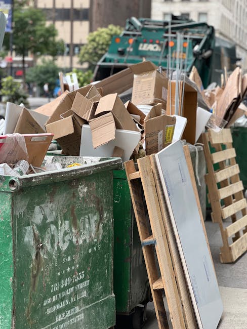 A large green waste container filled with various types of rubbish, including cardboard boxes, what appears to be packaging materials, and miscellaneous debris, situated outdoors on a street. The cardboard boxes are of different sizes, some flattened and some still in their box shape, with visible flaps and printed labels. Wooden pallets are leaning against the side of the container, with some damaged or partially disassembled. In the background, a refuse collection vehicle is visible, along with blurred elements of a city street, including trees, buildings, and parked cars. The scene suggests a site of on-site waste clearance or private rubbish disposal, with the waste ready for collection, possibly as part of a commercial or domestic rubbish removal process. The weather appears clear with natural daylight illuminating the scene, emphasizing the textures of the cardboard, wood, and the metallic surface of the waste container, supporting the context of professional rubbish collection services in an urban environment.